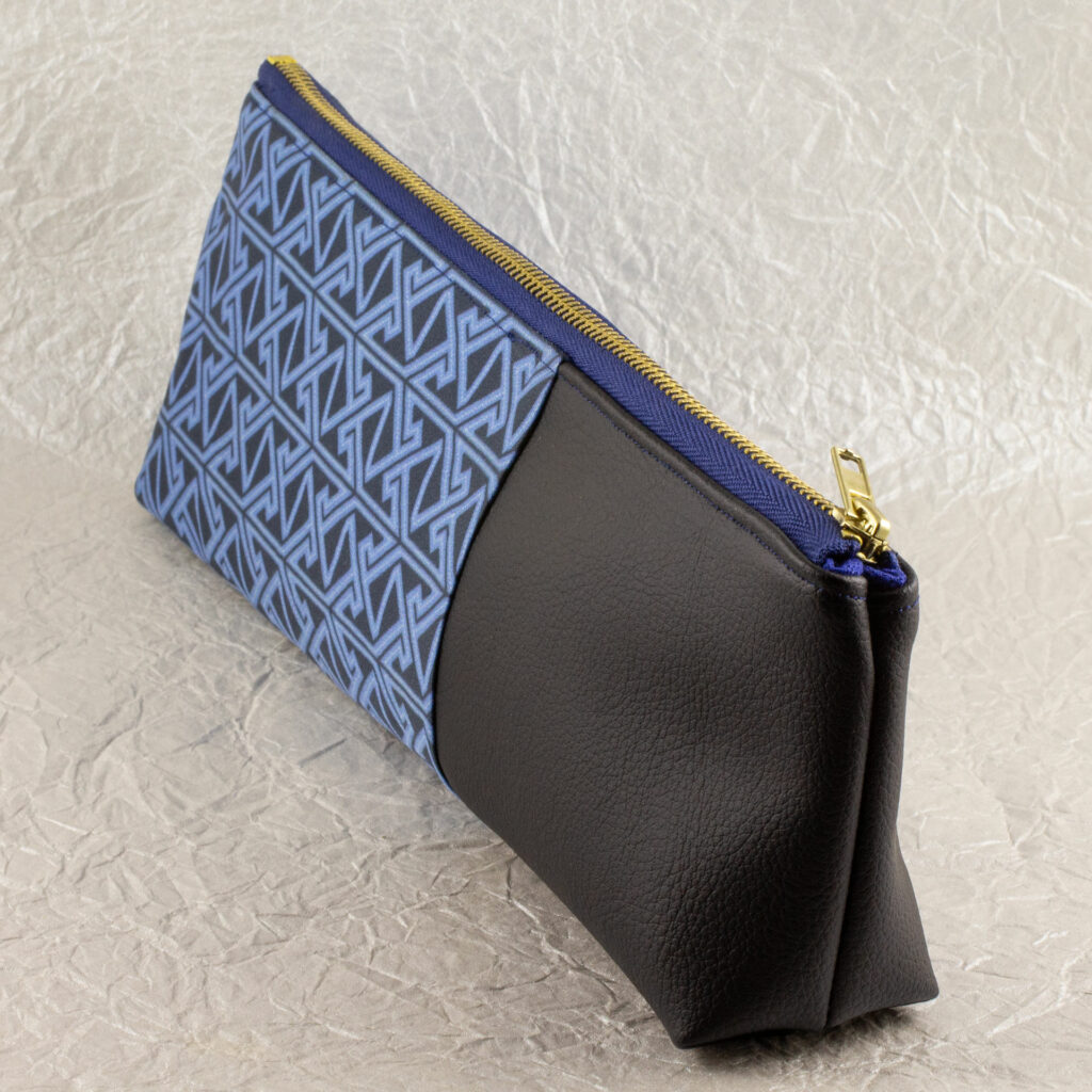 Three-quarter view of a pencil pouch that is about two-thirds fabric printed with a Celtic key pattern design in blue, and one-third black cactus leather. A blue and brass zipper closes the top. The surface and background are a pearlescent white crumpled-finish paper.