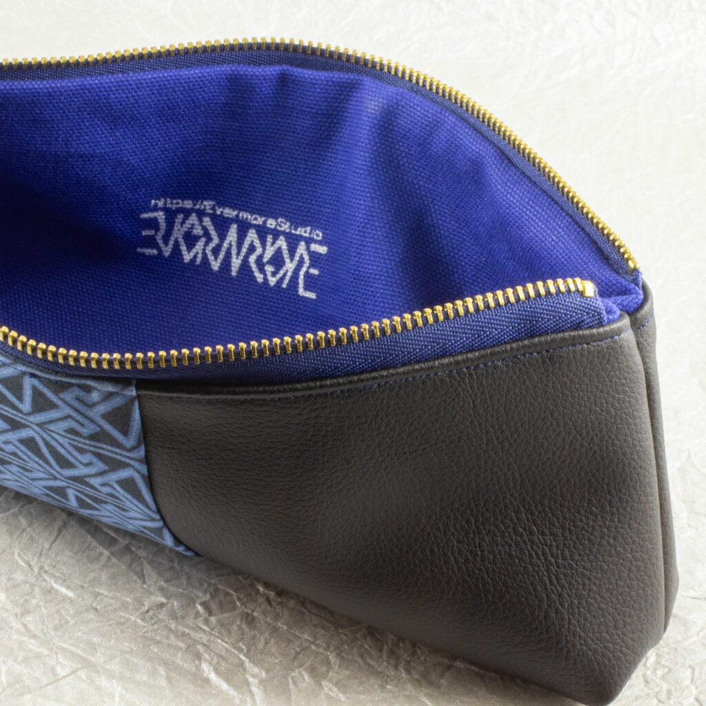 Close-up, front, top-down view of a pencil pouch that is about two-thirds fabric printed with a Celtic key pattern design in blue, and one-third black cactus leather. An open blue and brass zipper is at the top. The Evermore ambigram logo and web URL are visible printed in silver on the blue canvas lining. The surface and background are a pearlescent white crumpled-finish paper.