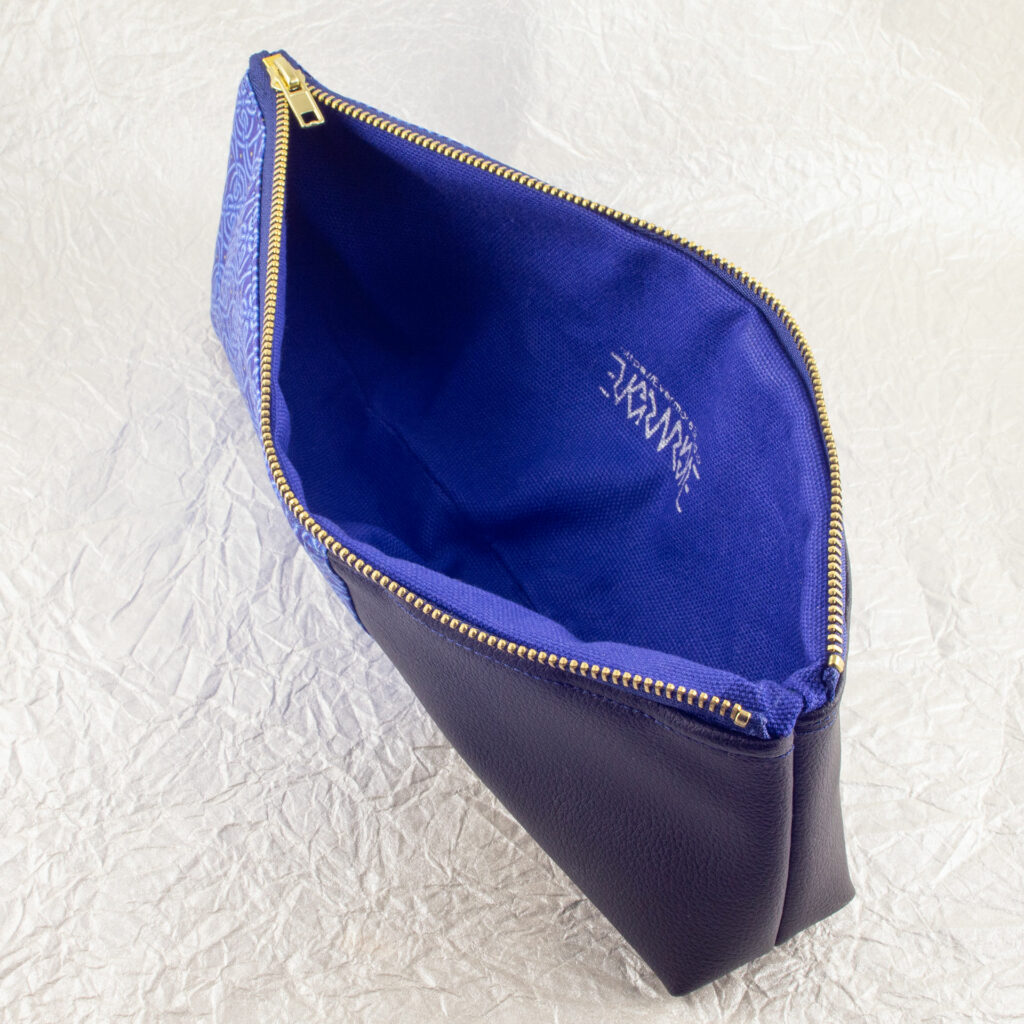 Top-down three-quarter view of a pencil pouch that is about two-thirds fabric printed with a continuous Celtic knot pattern design in blue, and one-third black cactus leather. An open blue and brass zipper is at the top. The Evermore ambigram logo and web URL are visible printed in silver on the blue canvas lining. The surface and background are a pearlescent white crumpled-finish paper.