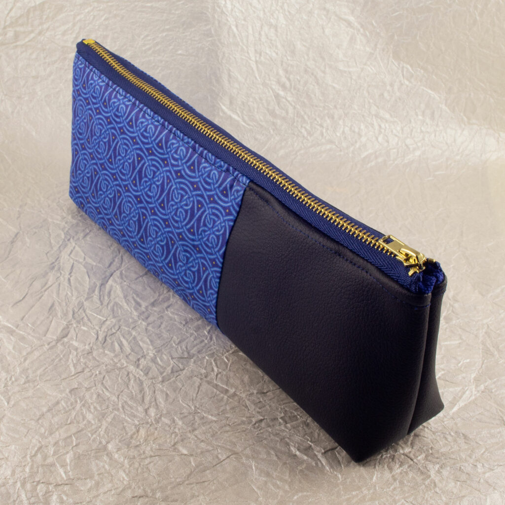 Three-quarter view of a pencil pouch that is about two-thirds fabric printed with a continuous Celtic knot pattern design in blue, and one-third black cactus leather. A blue and brass zipper closes the top. The surface and background are a pearlescent white crumpled-finish paper.