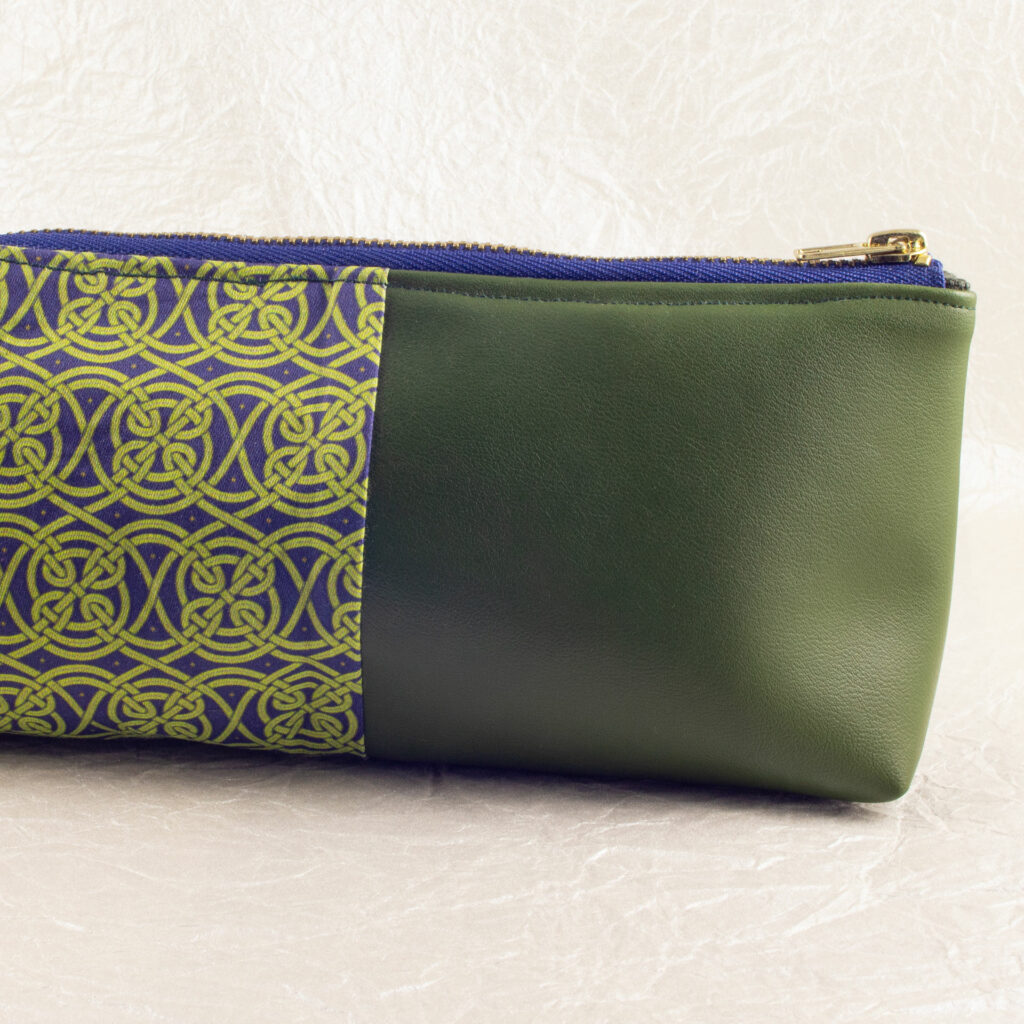 Close-up side view showing about half of a pencil pouch that is about two-thirds fabric printed with a continuous Celtic knot pattern design in green, and one-third green cactus leather.. A blue and brass zipper closes the top. The surface and background are a pearlescent white crumpled-finish paper.