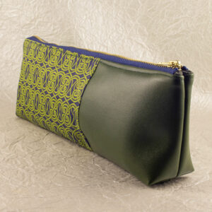 Three-quarter view of a pencil pouch that is about two-thirds fabric printed with a continuous Celtic knot pattern design in green, and one-third green cactus leather. A blue and brass zipper closes the top. The surface and background are a pearlescent white crumpled-finish paper.