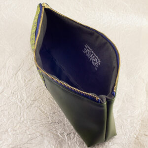 Top-down three-quarter view of a pencil pouch that is about two-thirds fabric printed with a Celtic key pattern design in green, and one-third green cactus leather. An open blue and brass zipper is at the top. The Evermore ambigram logo and web URL are visible printed in silver on the green canvas lining. The surface and background are a pearlescent white crumpled-finish paper.