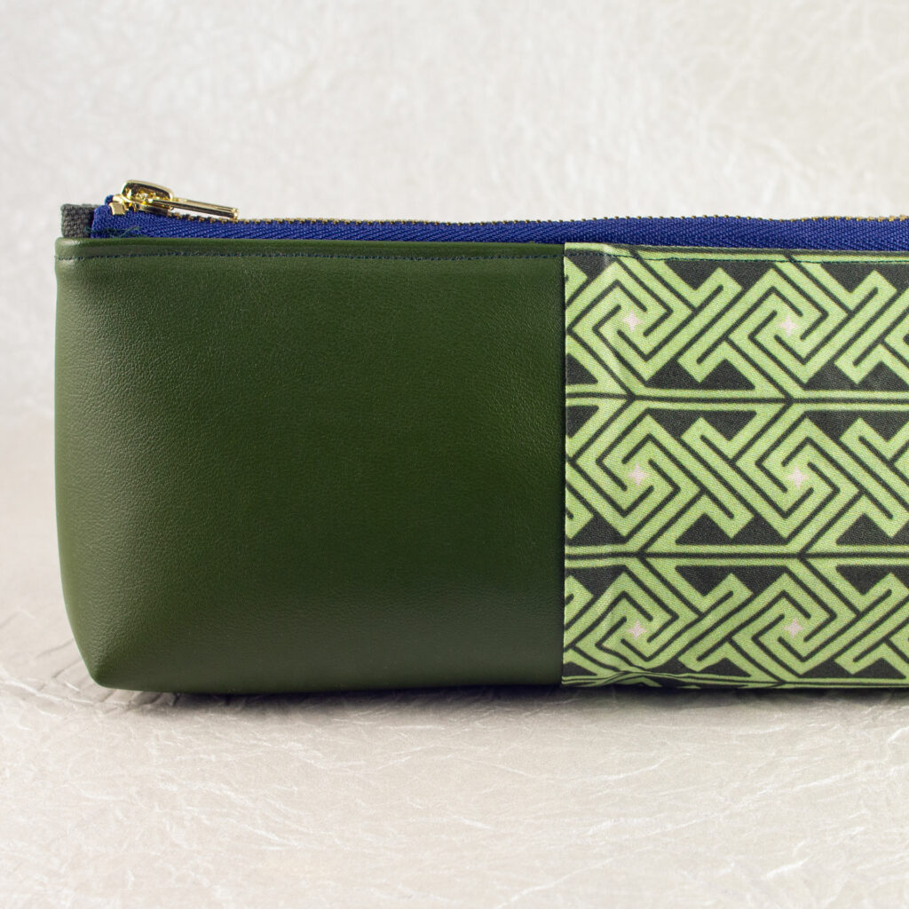 Close-up side view showing about half of a pencil pouch that is about two-thirds fabric printed with a Celtic key pattern design in green, and one-third green cactus leather. A blue and brass zipper closes the top. The surface and background are a pearlescent white crumpled-finish paper.