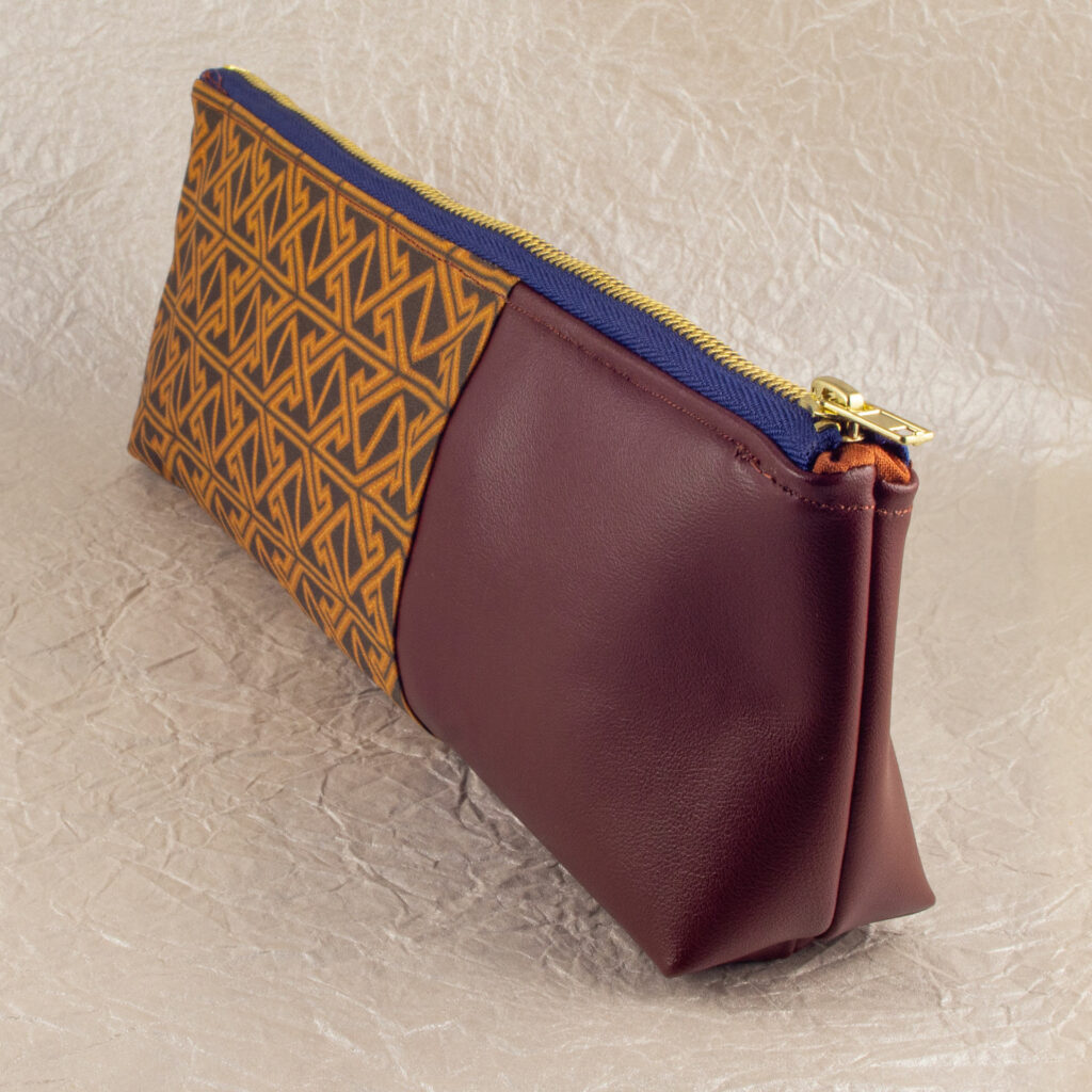 Three-quarter view of a pencil pouch that is about two-thirds fabric printed with a Celtic key pattern design in brown, and one-third red-brown cactus leather. A blue and brass zipper closes the top. The surface and background are a pearlescent white crumpled-finish paper.