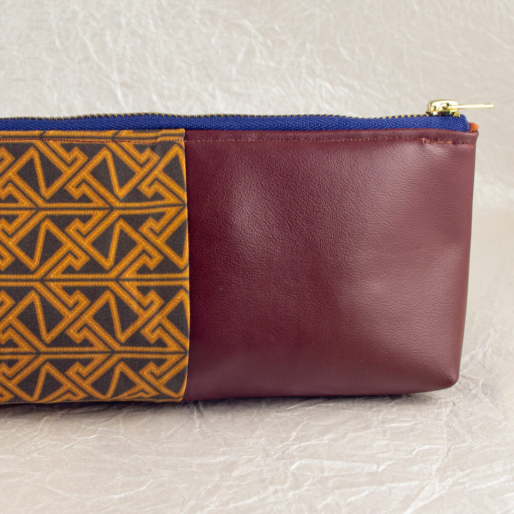 Close-up side view showing about half of a pencil pouch that is about two-thirds fabric printed with a Celtic key pattern design in brown, and one-third red-brown cactus leather. A blue and brass zipper closes the top. The surface and background are a pearlescent white crumpled-finish paper.