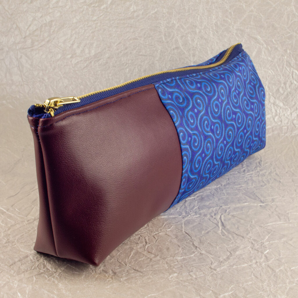Three-quarter view of a pencil pouch that is about two-thirds fabric printed with a Celtic spirals design in blue, and one-third red-brown cactus leather. A blue and brass zipper closes the top. The surface and background are a pearlescent white crumpled-finish paper.