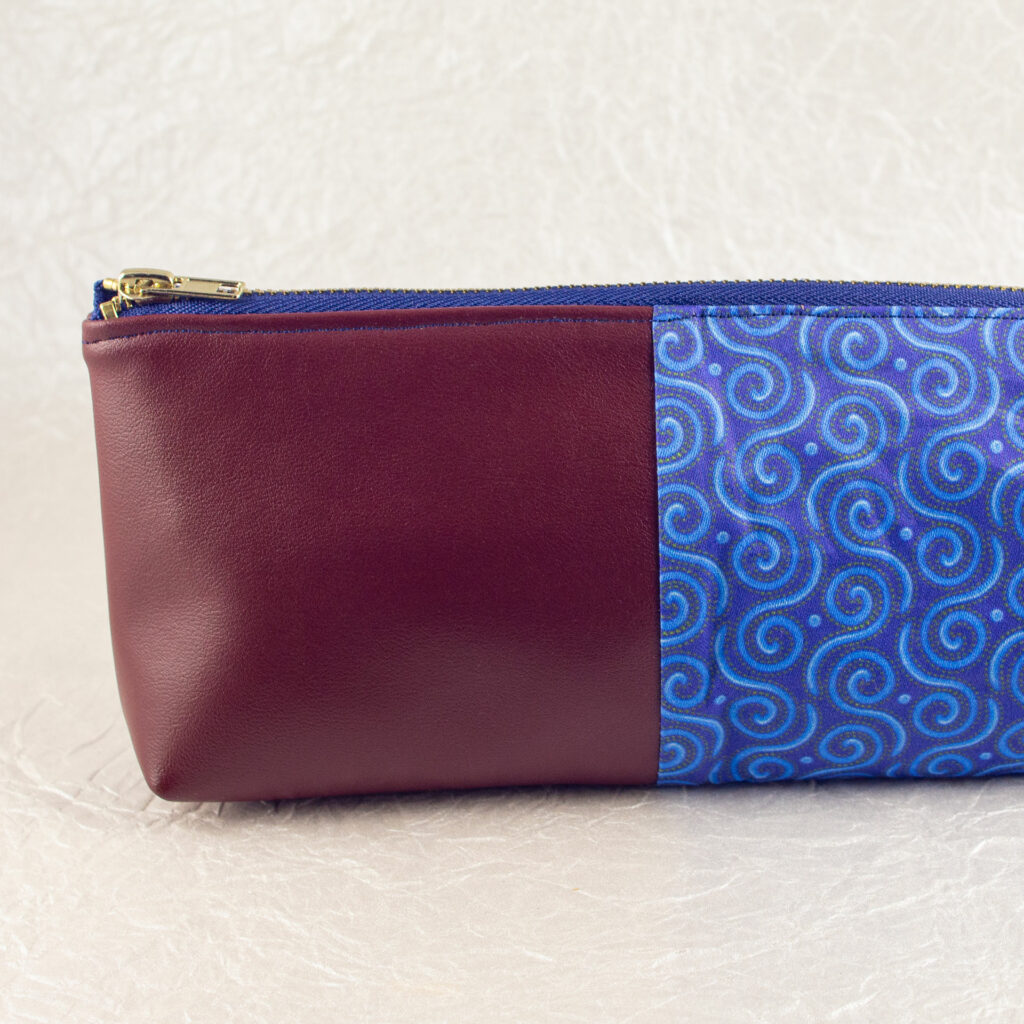 Close-up side view showing about half of a pencil pouch that is about two-thirds fabric printed with a Celtic spirals design in blue, and one-third red-brown cactus leather. A blue and brass zipper closes the top. The surface and background are a pearlescent white crumpled-finish paper.