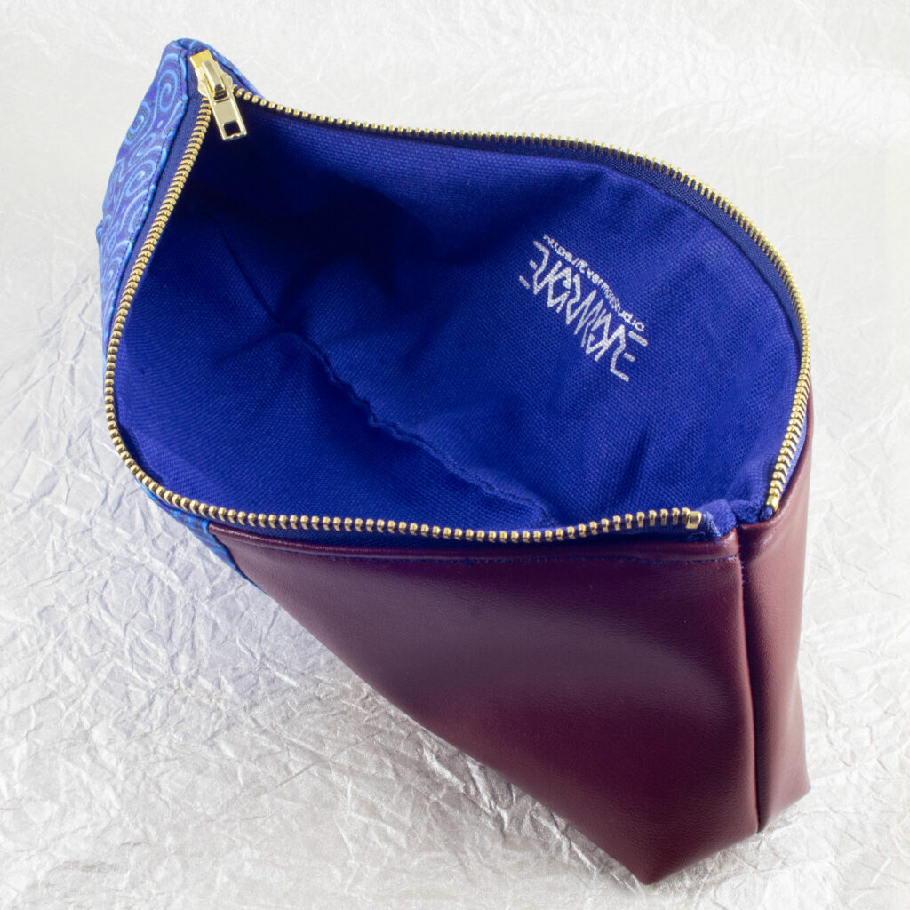 Top-down three-quarter view of a pencil pouch that is about two-thirds fabric printed with a Celtic spirals design in blue, and one-third red-brown cactus leather. An open blue and brass zipper is at the top. The Evermore ambigram logo and web URL are visible printed in silver on the blue canvas lining. The surface and background are a pearlescent white crumpled-finish paper.