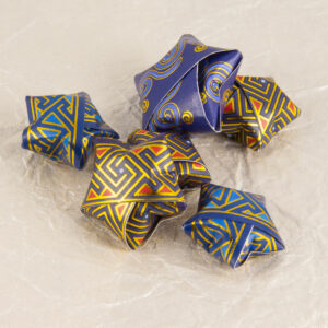 A number of small origami stars made from Celtic art washi tape. Three use my Squared Spirals key pattern in metallic gold, deep purple, and red. Two use my first key pattern washi tape in shades of blue and metallic gold. One slightly larger star is blue with lighter blue spirals with metallic gold accents. They are scattered on a background of off-white crinkled (momigami) paper.