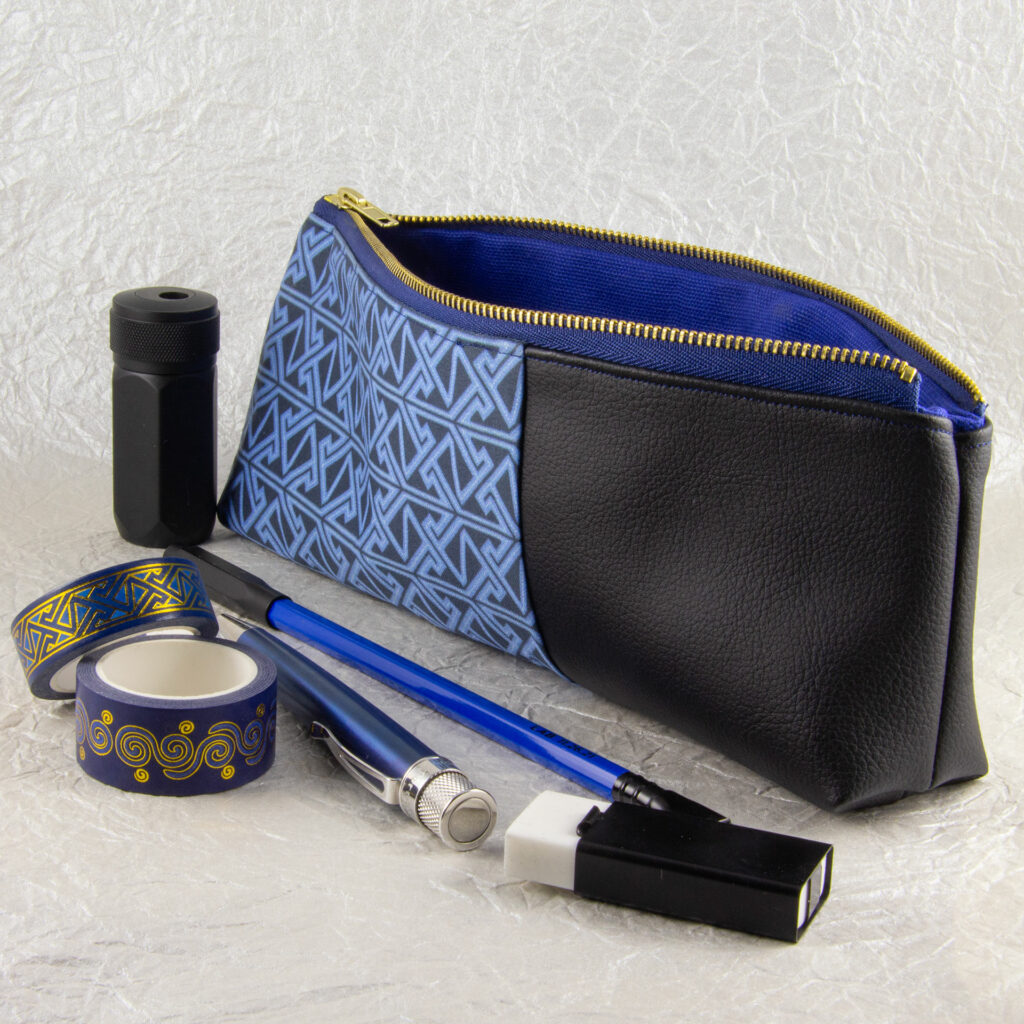 Three-quarter view of a pencil pouch that is about two-thirds fabric printed with a continuous Celtic key pattern design in blue, and one-third black cactus leather. An open blue and brass zipper is at the top. A blue Blackwing pencil with a black point guard, a metallic blue Retro 51 pen, a rectangular white eraser in a black metal sheath, a cylindrical black metal pencil sharpener, and two rolls of different Celtic art washi tape sit in front of the pouch. The surface and background are a pearlescent white crumpled-finish paper.