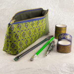 Three-quarter view of a pencil pouch that is about two-thirds fabric printed with a continuous Celtic knotwork design in green, and one-third green cactus leather. An open blue and brass zipper is at the top. A Blackwing pencil with a point guard, a brighter green pencil, a Retro 51 Cioppino pen, and three rolls of different Celtic art washi tape sit in front of the pouch. The surface and background are a pearlescent white crumpled-finish paper.