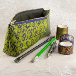 Three-quarter view of a pencil pouch that is about two-thirds fabric printed with a continuous Celtic knotwork design in green, and one-third green cactus leather. An open blue and brass zipper is at the top. A Blackwing pencil with a point guard, a brighter green pencil, a Retro 51 Cioppino pen, and three rolls of different Celtic art washi tape sit in front of the pouch. The surface and background are a pearlescent white crumpled-finish paper.