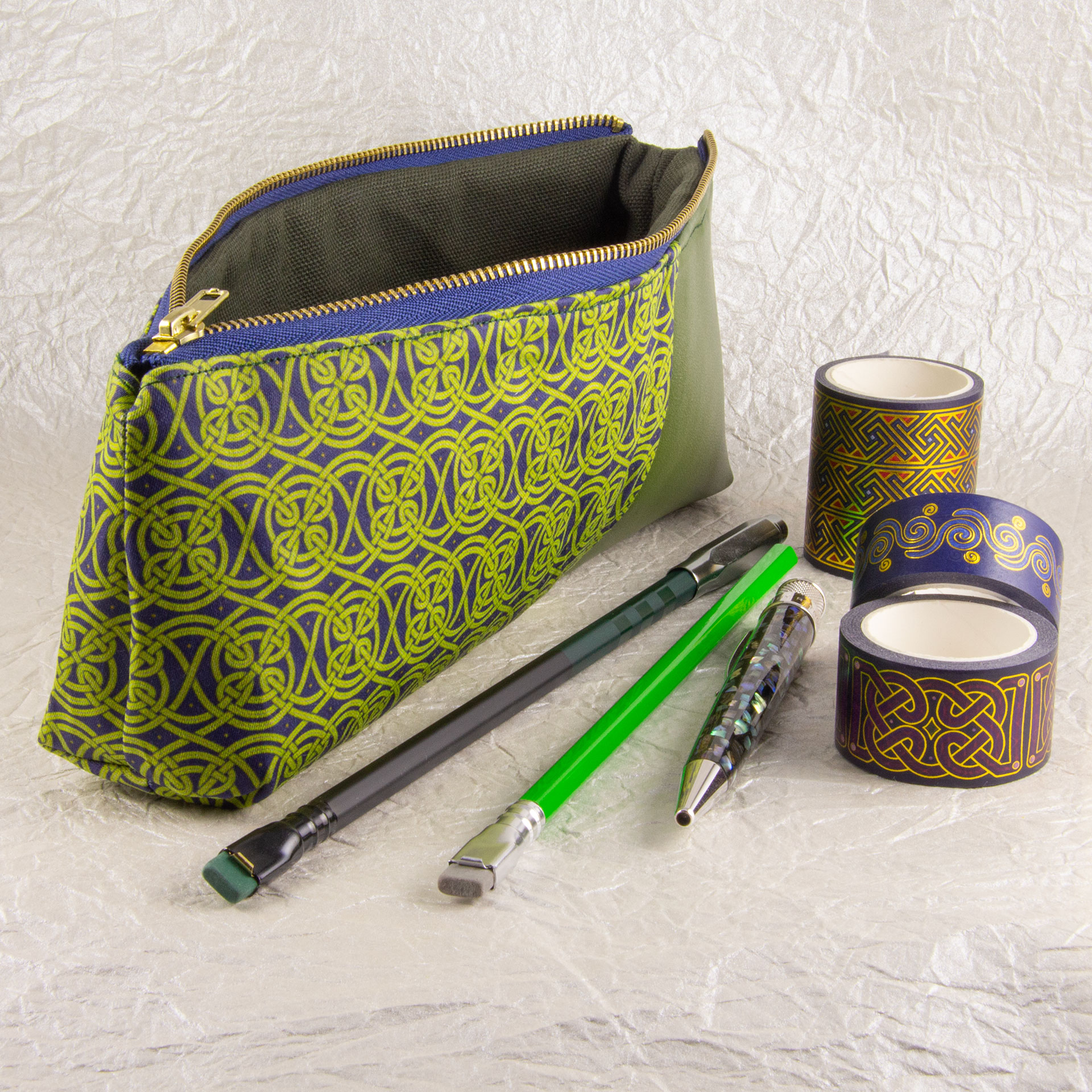 Three-quarter view of a pencil pouch that is about two-thirds fabric printed with a continuous Celtic knotwork design in green, and one-third green cactus leather. An open blue and brass zipper is at the top. A Blackwing pencil with a point guard, a brighter green pencil, a Retro 51 Cioppino pen, and three rolls of different Celtic art washi tape sit in front of the pouch. The surface and background are a pearlescent white crumpled-finish paper.