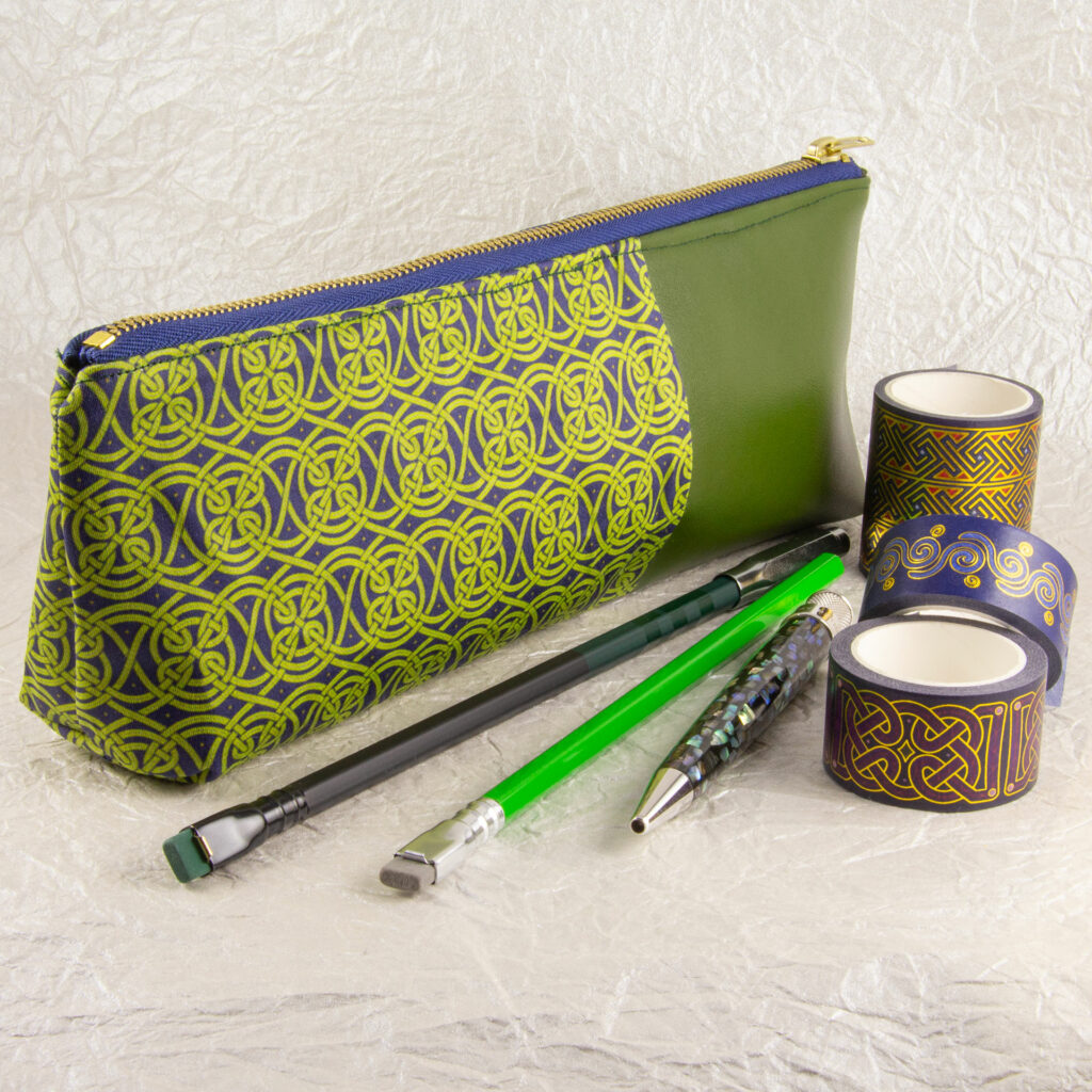 Three-quarter view of a pencil pouch that is about two-thirds fabric printed with a continuous Celtic knotwork design in green, and one-third green cactus leather. A blue and brass zipper closes the top. A Blackwing pencil with a point guard, a brighter green pencil, a Retro 51 Cioppino pen, and three rolls of different Celtic art washi tape sit in front of the pouch. The surface and background are a pearlescent white crumpled-finish paper.