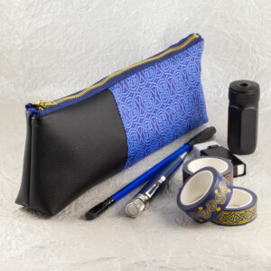 Three-quarter view of a pencil pouch that is about two-thirds fabric printed with a continuous Celtic knot pattern design in blue, and one-third black cactus leather. A blue and brass zipper closes the top. A blue Blackwing pencil with a black point guard, a metallic blue Retro 51 pen, a rectangular white eraser in a black metal sheath, a cylindrical black metal pencil sharpener, and three rolls of different Celtic art washi tape sit in front of the pouch. The surface and background are a pearlescent white crumpled-finish paper.