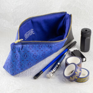 Three-quarter view of a pencil pouch that is about two-thirds fabric printed with a continuous Celtic knot pattern design in blue, and one-third black cactus leather. An open blue and brass zipper at the top reveals a blue canvas liner with the Evermore ambigram and Evermore Studio URL printed in silver. A blue Blackwing pencil with a black point guard, a metallic blue Retro 51 pen, a rectangular white eraser in a black metal sheath, a cylindrical black metal pencil sharpener, and three rolls of different Celtic art washi tape sit in front of the pouch. The surface and background are a pearlescent white crumpled-finish paper.