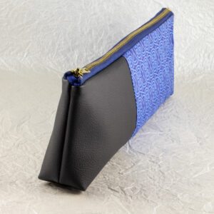 Three-quarter view of a pencil pouch that is about two-thirds fabric printed with a continuous Celtic knot pattern design in blue, and one-third black cactus leather. A blue and brass zipper closes the top. Seen from the black cactus leather end. The surface and background are a pearlescent white crumpled-finish paper.