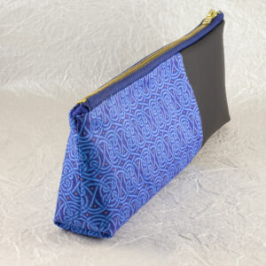 Three-quarter view of a pencil pouch that is about two-thirds fabric printed with a continuous Celtic knot pattern design in blue, and one-third black cactus leather. A blue and brass zipper closes the top. Seen from the knotwork fabric end. The surface and background are a pearlescent white crumpled-finish paper.