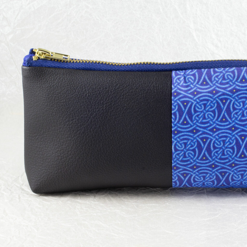 Partial side view of a pencil pouch that is about two-thirds fabric printed with a continuous Celtic knot pattern design in blue, and one-third black cactus leather. A blue and brass zipper closes the top. Image shows the black cactus leather end with a little bit of the fabric. The surface and background are a pearlescent white crumpled-finish paper.
