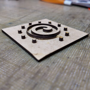 A wooden embossing plate made from pieces of MDF glued to a base: a Celtic spiral surrounded by 11 small circles. One circle is missing.