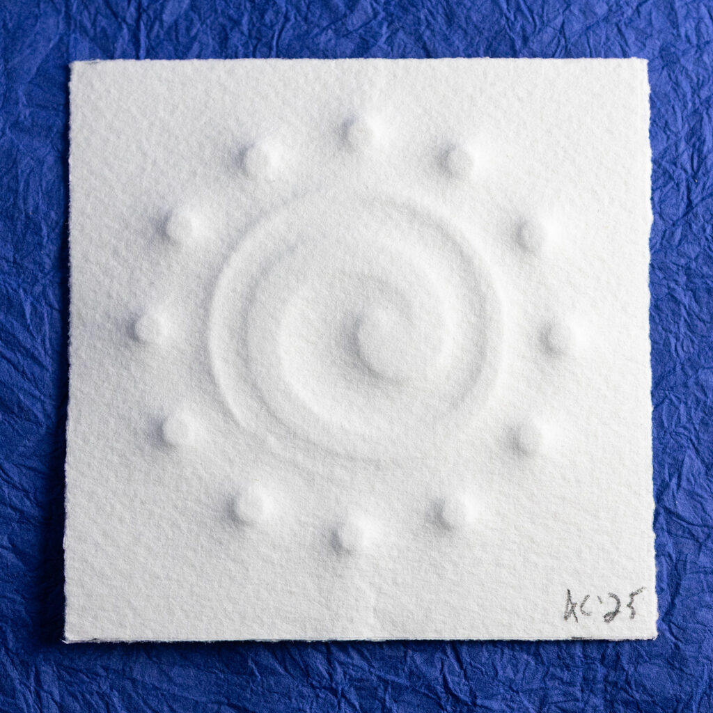 An off-center counter-clockwise tapering spiral surrounded by twelve small circles is embossed on a small (70mm) square of toothy paper. It sits on a background of crinkled blue paper (momigami).