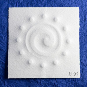An off-center counter-clockwise tapering spiral surrounded by twelve small circles is embossed on a small (70mm) square of toothy paper. It sits on a background of crinkled blue paper (momigami).