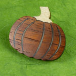 A small pin in the shape of a pumpkin made from orangey padauk with ribs of inlaid walnut and a white oak stem resting on a background of green paper with fiber inclusions.