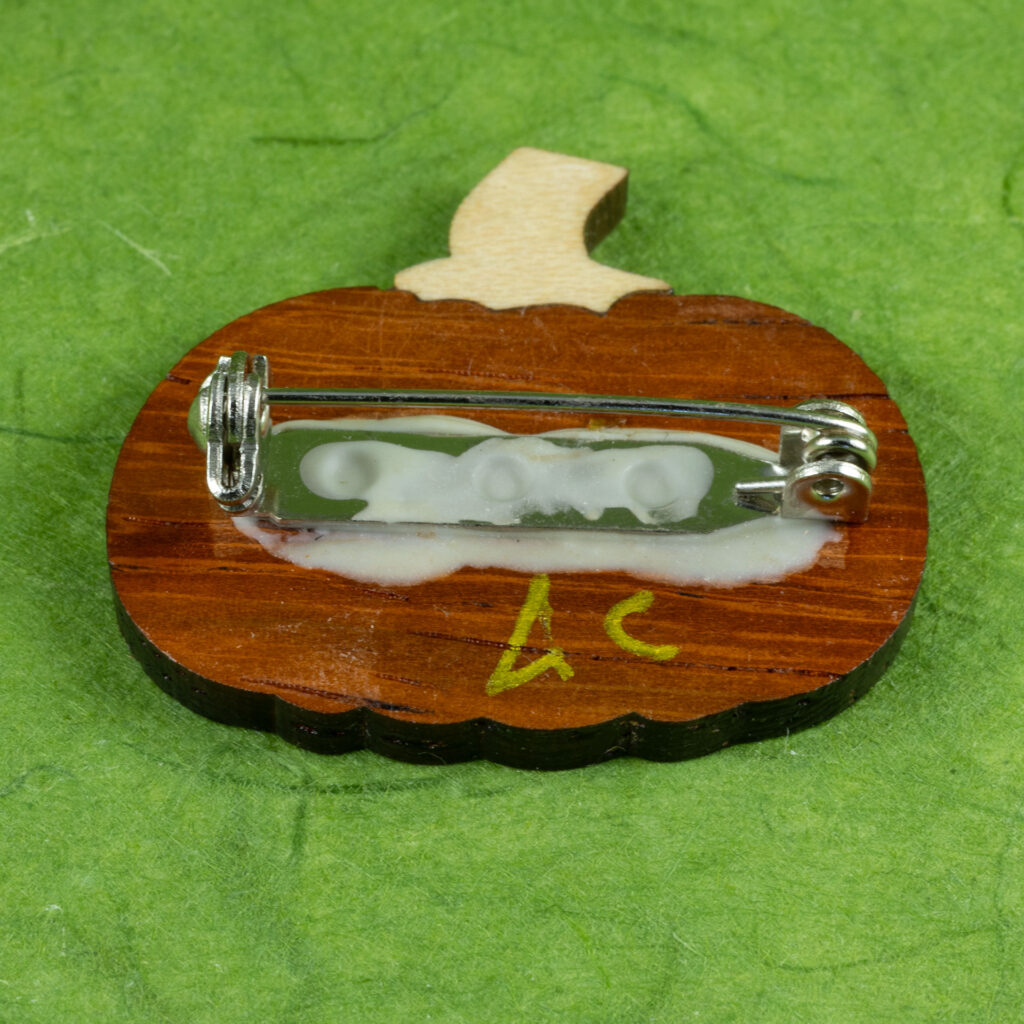 Back side of the wood inlay pumpkin pin showing the glued-on pin backing and a metallic gold AC inscribed as a maker's mark.