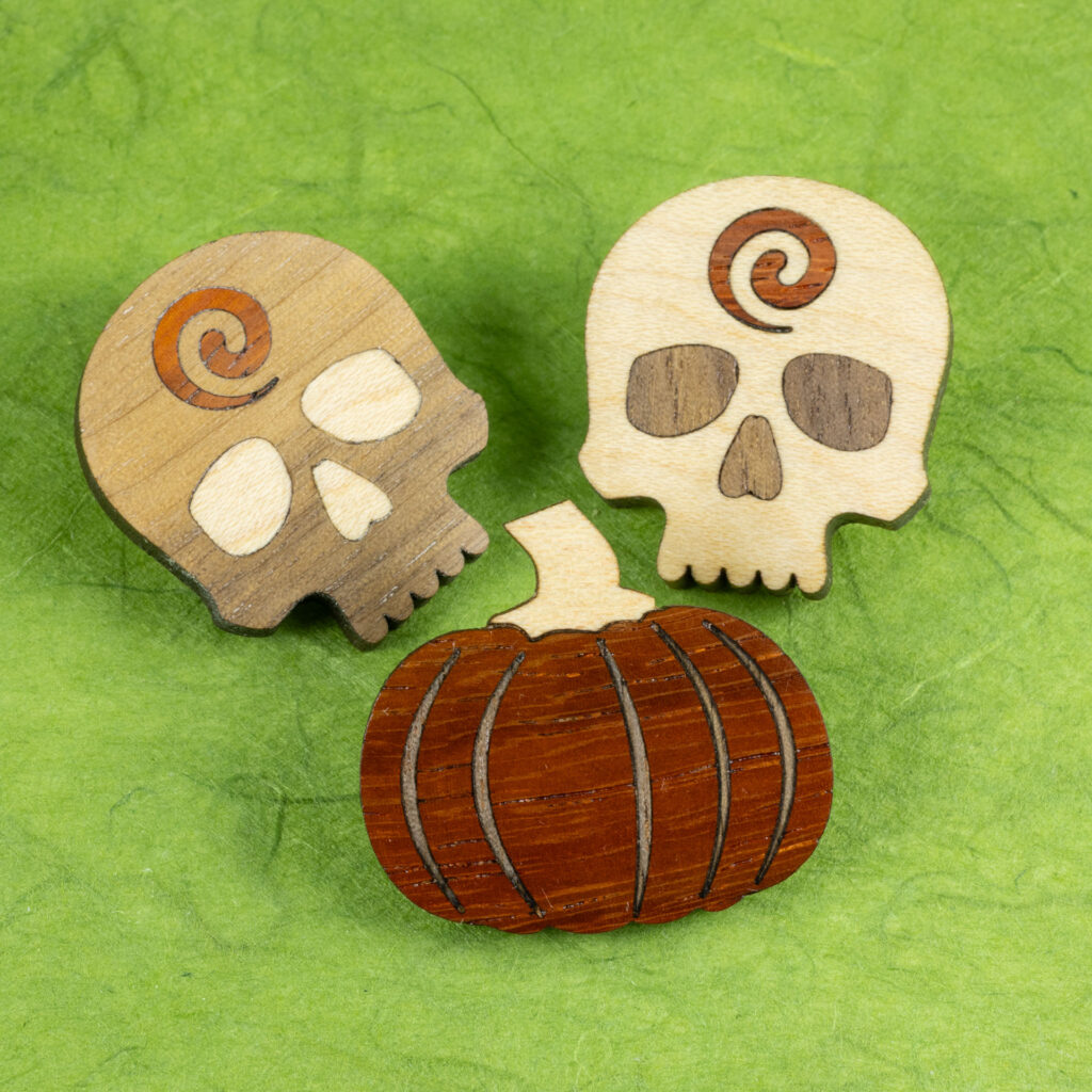 A trio of small wood inlay pins: two skulls from white oak, walnut and, padauk (one predominantly white oak, the other predominantly walnut), and a pumpkin pin of orange padauk inlaid with walnut ribs and a white oak stem.