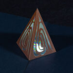 A tall, thin metallic copper four-sided pyramid with curvy organic cut outs on each face sits on a slightly pearlescent dark blue surface. The pyramid is lit from the inside, visible through the cut outs, with the light diffracted into rainbow shards. Three-quarter view from a little above, highlighting one face of the pyramid.