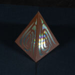 A tall, thin metallic copper four-sided pyramid with curvy organic cut outs on each face sits on a slightly pearlescent dark blue surface. The pyramid is lit from the inside, visible through the cut outs, with the light diffracted into rainbow shards. Three-quarter view from above towards one edge of the pyramid.