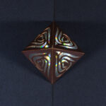 A tall, thin metallic copper four-sided pyramid with curvy organic cut outs on each face sits on a slightly pearlescent dark blue surface. The pyramid is lit from the inside, visible through the cut outs, with the light diffracted into rainbow shards. Top-down view from directly overhead showing all four faces of the pyramid.