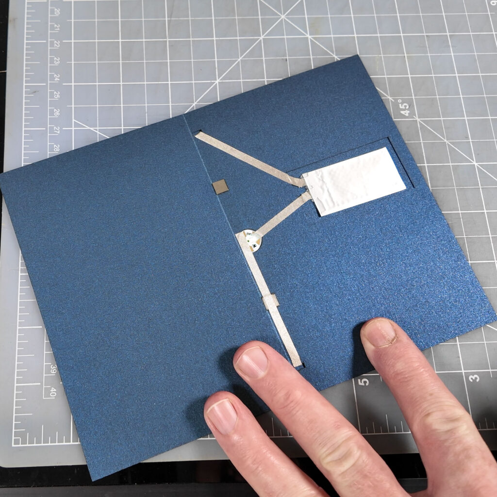 The open dark blue card prior to installing the pyramid sits on a translucent cutting mat with a ruled grid. A hand holds it open. The interior portion of the circuit is visible, including traces made from silvery conductive fabric, a flat battery, and a heart-shaped Circuit Sticker.