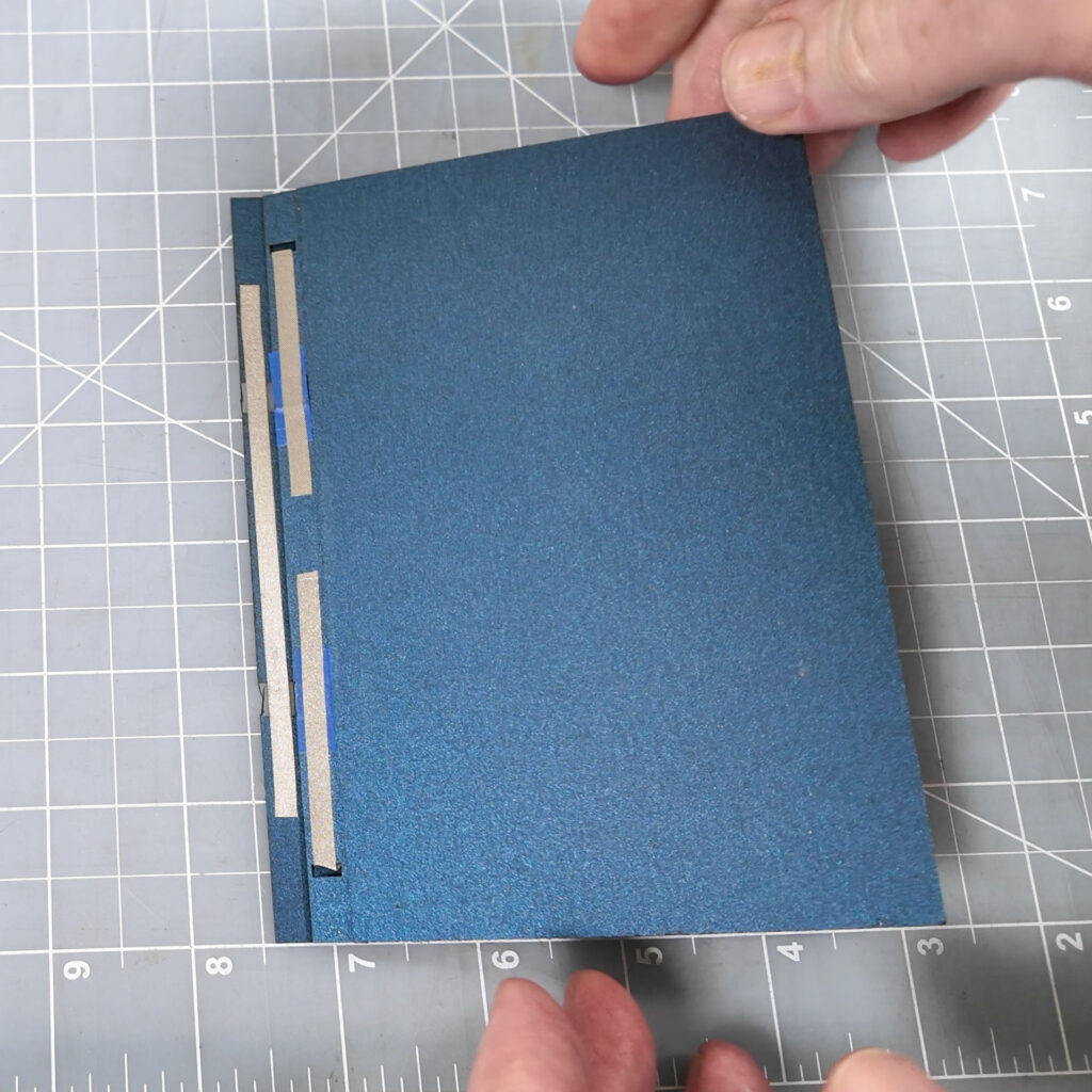 The closed dark blue card sitting on a translucent cutting mat with a ruled grid. Hands appear to have just set the card down. On the left edge of the back of the card can be seen a strip of grey conductive fabric tape with two similar strips with a gap on the other side of the hinge.
