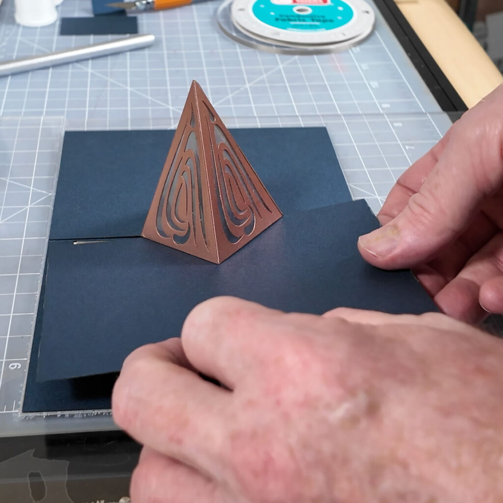 The last panel is being added to a dark blue card with a metallic copper pyramid in the center. The pyramid has curvy organic cut outs in the faces. The card is sitting on a translucent cutting mat with a ruled grid. One hand holds the card while another pulls a mounting thread through a hole in the final panel.
