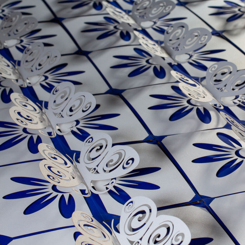 A collection of somewhat stylized kirigami pop up pearlescent white paper butterfly cards arranged side-by-side. The butterflies have Celtic spiral designs cut into the wingss. There is a flower design cut into the base, which allows the cobalt blue backing card to show through for the petals. Above and almost head-on view.