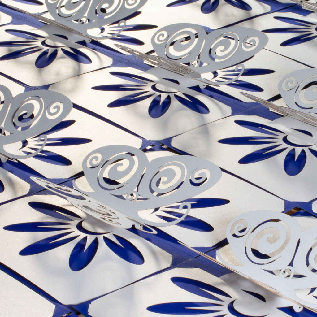 A collection of somewhat stylized kirigami pop up pearlescent white paper butterfly cards arranged side-by-side. The butterflies have Celtic spiral designs cut into the wingss. There is a flower design cut into the base, which allows the cobalt blue backing card to show through for the petals. Three-quarters above left view.
