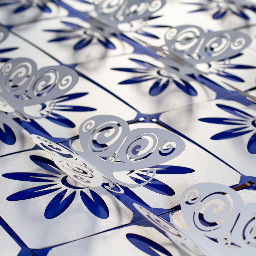 A collection of somewhat stylized kirigami pop up pearlescent white paper butterfly cards arranged side-by-side. The butterflies have Celtic spiral designs cut into the wingss. There is a flower design cut into the base, which allows the cobalt blue backing card to show through for the petals. Three-quarters above left view.
