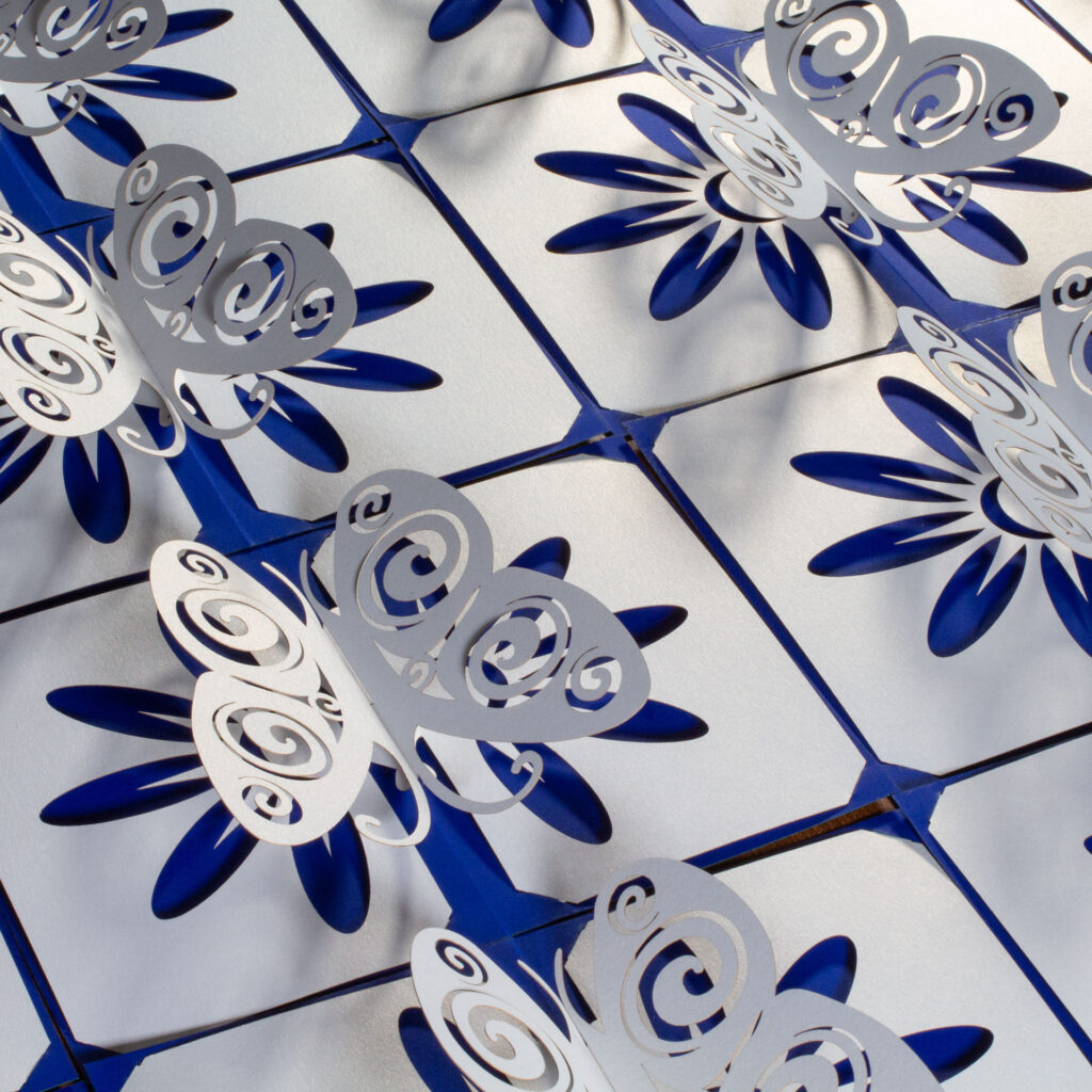 A collection of somewhat stylized kirigami pop up pearlescent white paper butterfly cards arranged side-by-side. The butterflies have Celtic spiral designs cut into the wingss. There is a flower design cut into the base, which allows the cobalt blue backing card to show through for the petals. Three-quarters above left view.
