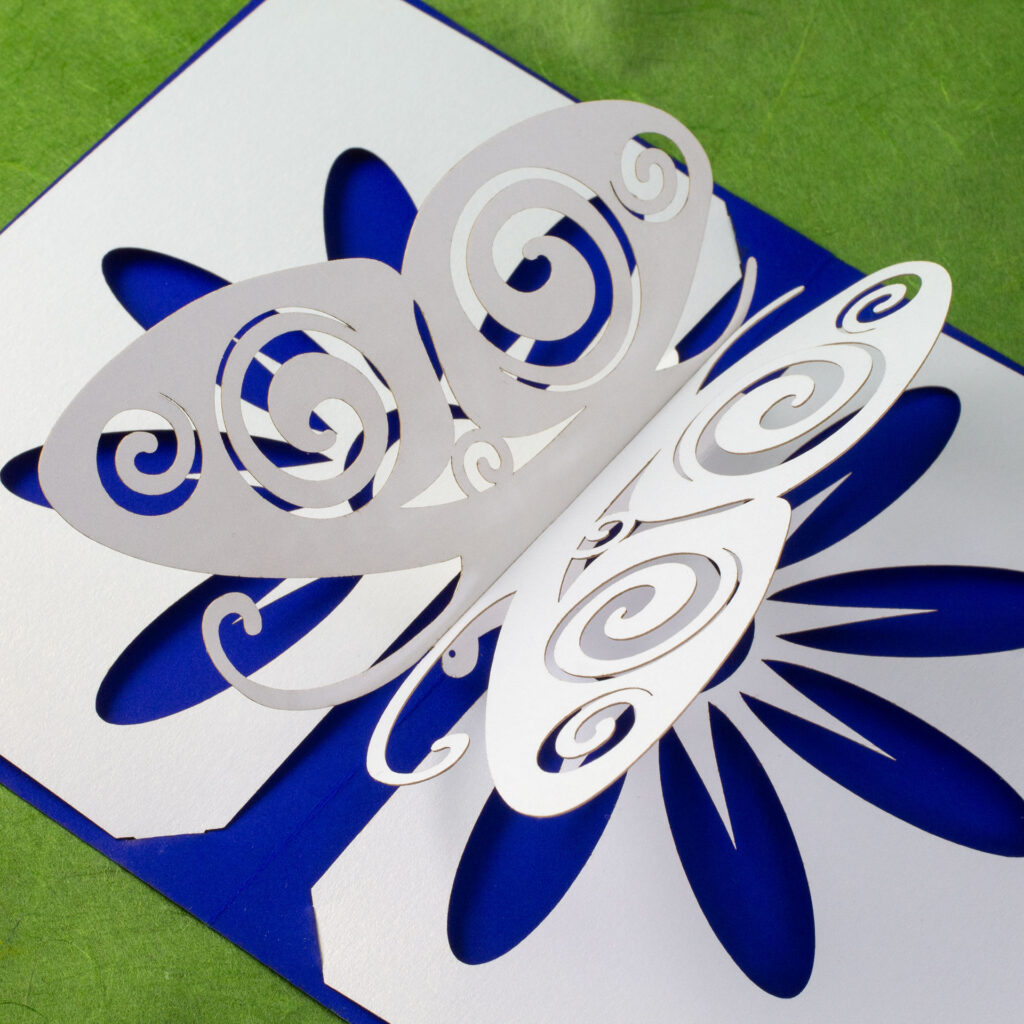 Somewhat stylized kirigami pop up pearlescent white paper butterfly with Celtic spiral designs cut into the wings. There is a flower design cut into the base, which allows the cobalt blue backing card to show through for the petals. Three-quarters above right view (higher up).