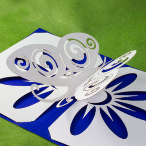 Somewhat stylized kirigami pop up pearlescent white paper butterfly with Celtic spiral designs cut into the wings. There is a flower design cut into the base, which allows the cobalt blue backing card to show through for the petals. Three-quarters above right view.