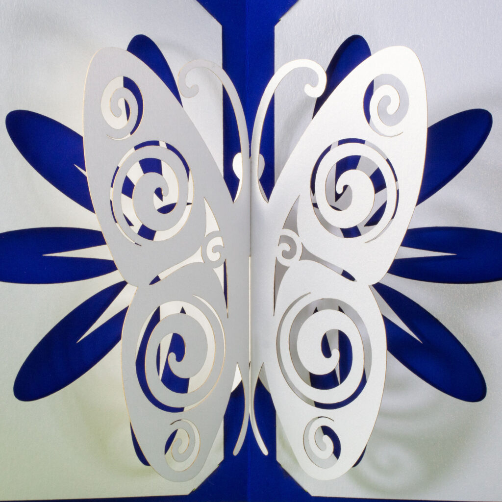 Somewhat stylized kirigami pop up pearlescent white paper butterfly with Celtic spiral designs cut into the wings. There is a flower design cut into the base, which allows the cobalt blue backing card to show through for the petals. Top-down view.