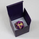 A 2.5"/63mm cubic purple box sits on a pearlescent white surface. A Celtic hatch-filled knotwork design adorns the visible face. The top is open. Inside a purple and yellow 20-sided die sits on a silver-colored paper insert with an intricately cut lattice that allows it to conform to the shape of the die.