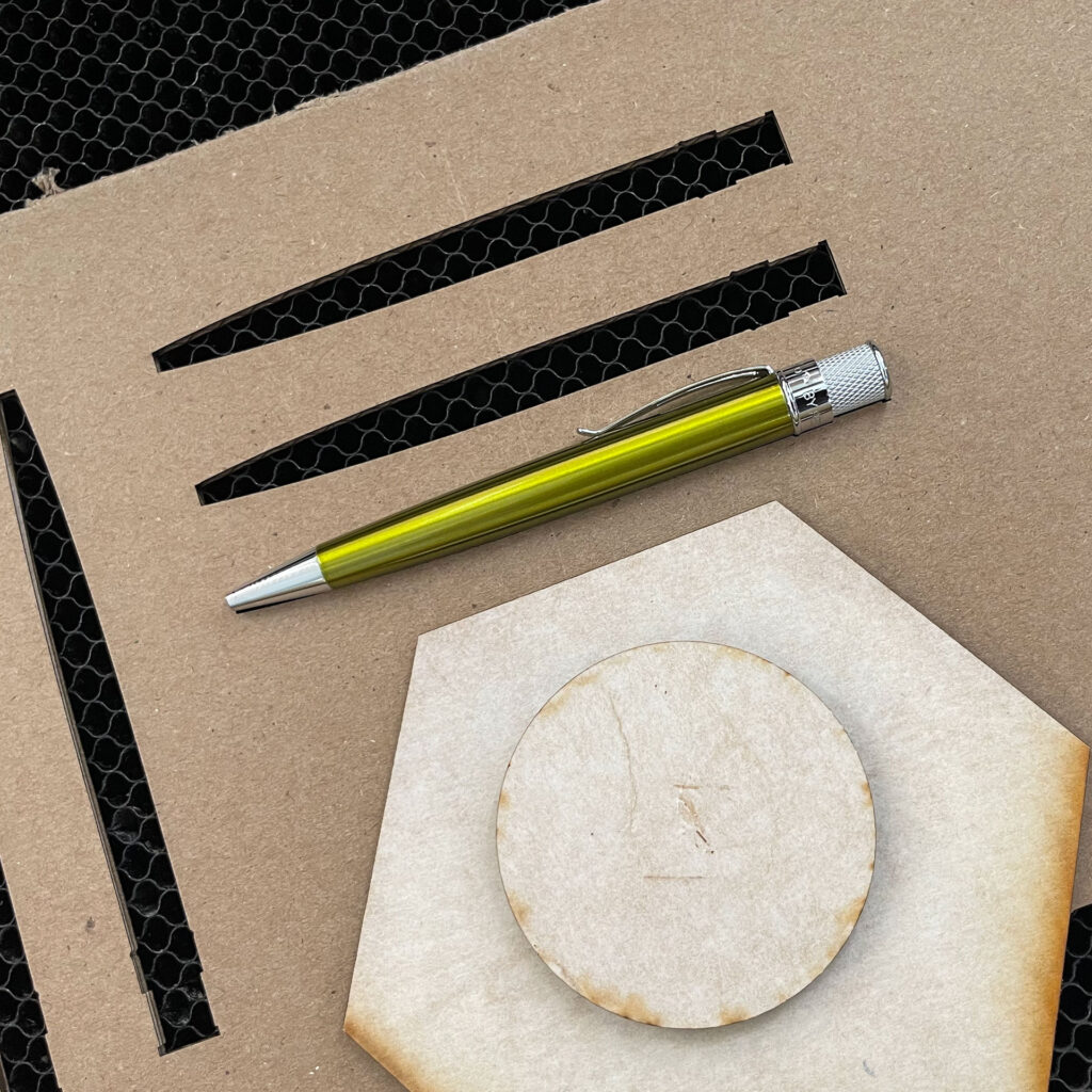 Cardboard jig with pen-shaped cutouts, one contains a metallic avocado green Retro 51 Tornado pen. A circular piece stacked on a hexagonal piece sits on top of the jig to serve as a focus point.