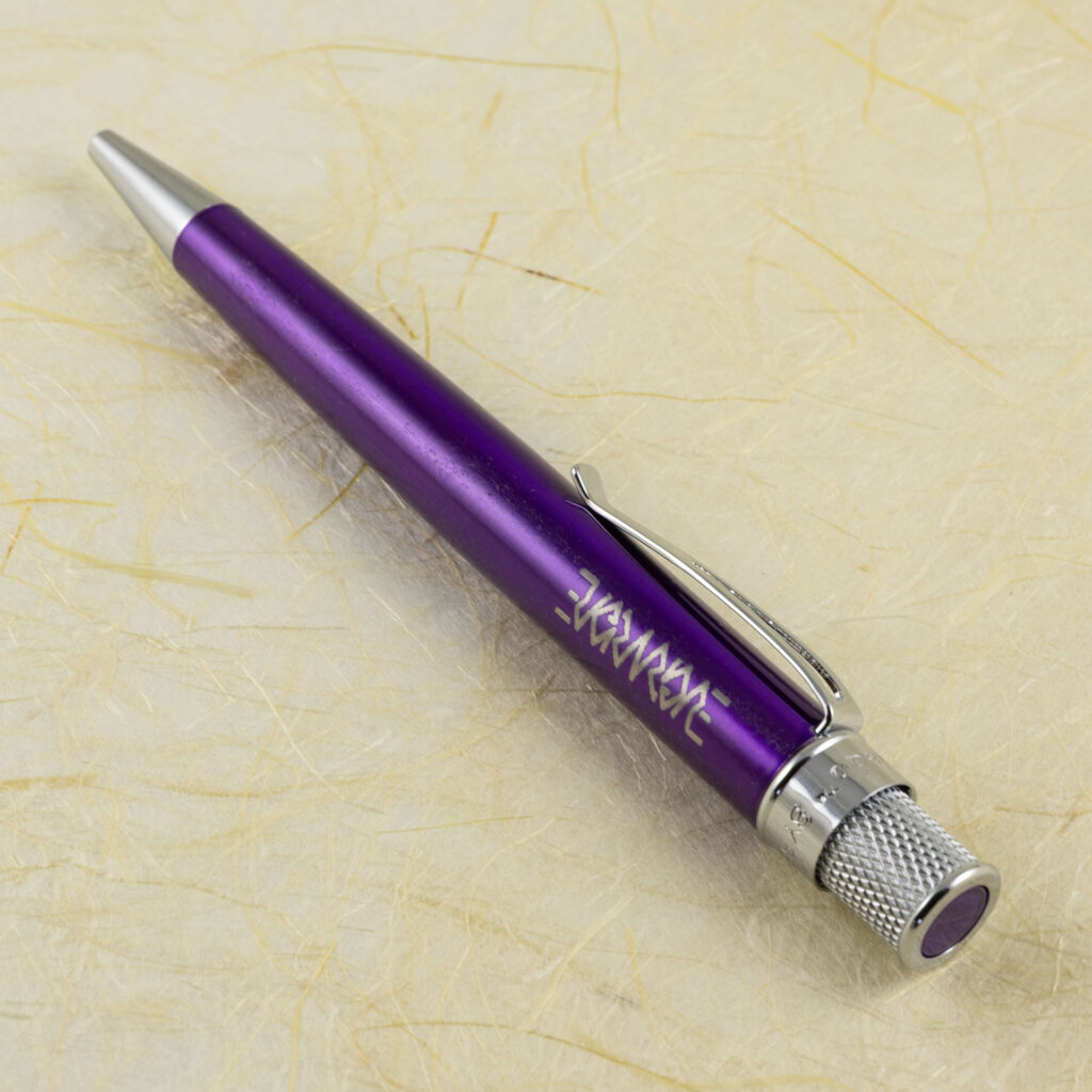 A metal pen lacquered in purple and engraved with the Evermore ambigram lies diagonally on a surface of fibrous off-white handmade paper. The wide end of the pen in the lower left is knurled and has a dot of the purple lacquer on the end.