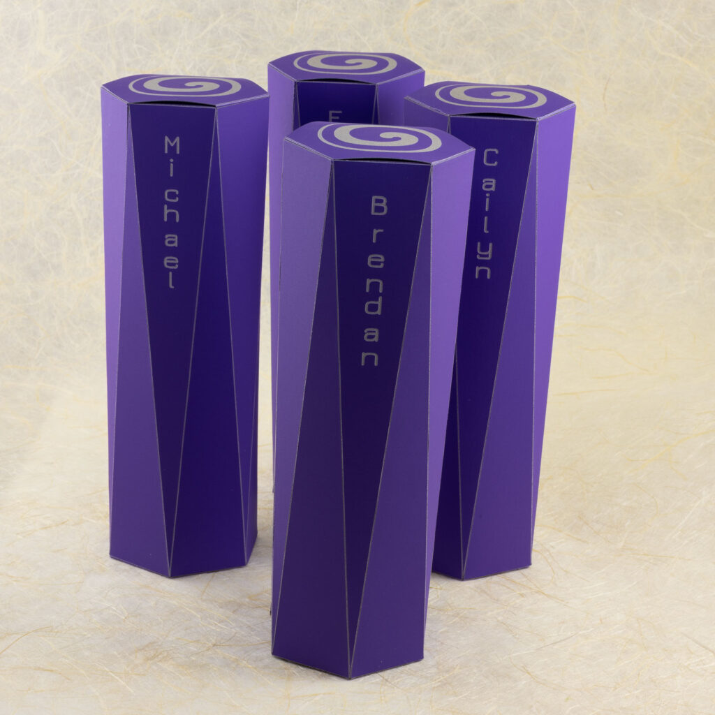 Four long purple boxes. The ends are hexagonal. The sides extending from each hexagonal face are long triangles that come to a point at the vertices of the hexagon on the opposite end. The top hexagonal face is engraved with an off-center spiral in light grey. There are names engraved on the front faces: Michael, Brendan, Cailyn, and Evie. View from the front with the boxes standing upright.