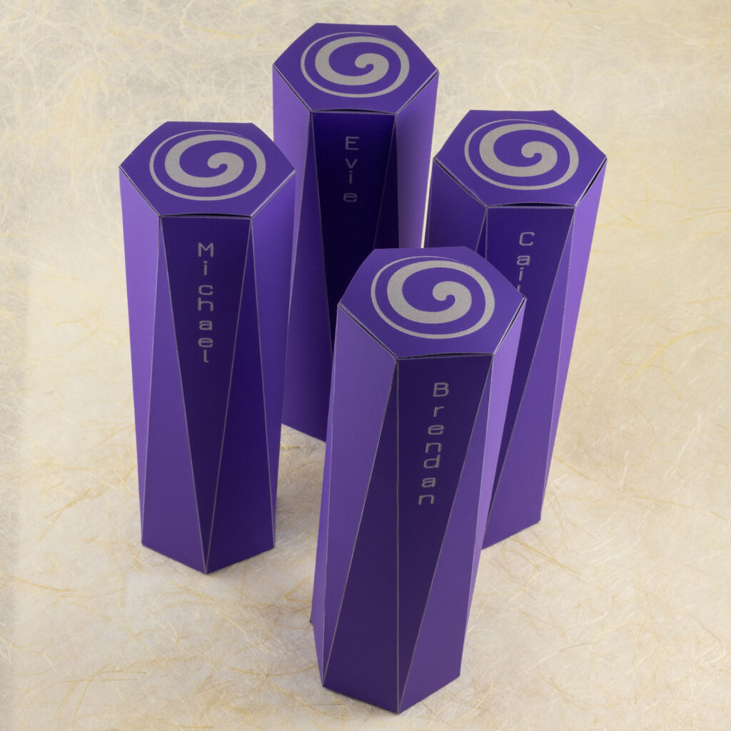 Four long purple boxes. The ends are hexagonal. The sides extending from each hexagonal face are long triangles that come to a point at the vertices of the hexagon on the opposite end. The top hexagonal face is engraved with an off-center spiral in light grey. There are names engraved on the front faces: Michael, Brendan, Cailyn, and Evie. View from above-front, looking down, with the boxes standing upright.