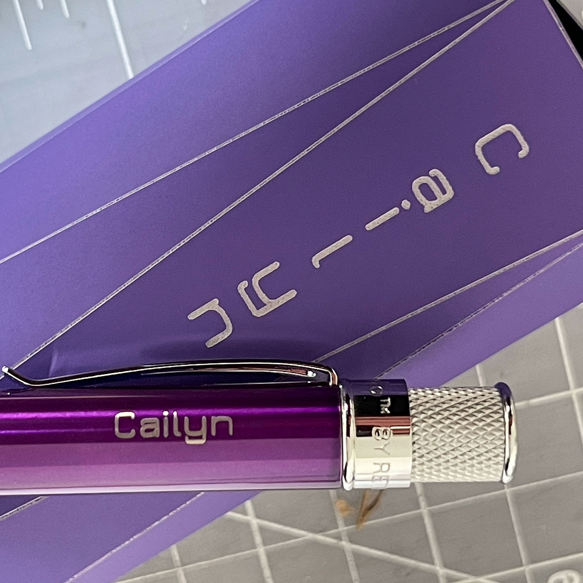 Close-up of the end of a purple Retro 51 Classic Lacquer pen engraved with the name Cailyn, lying on top of a purple box engraved with the same name.