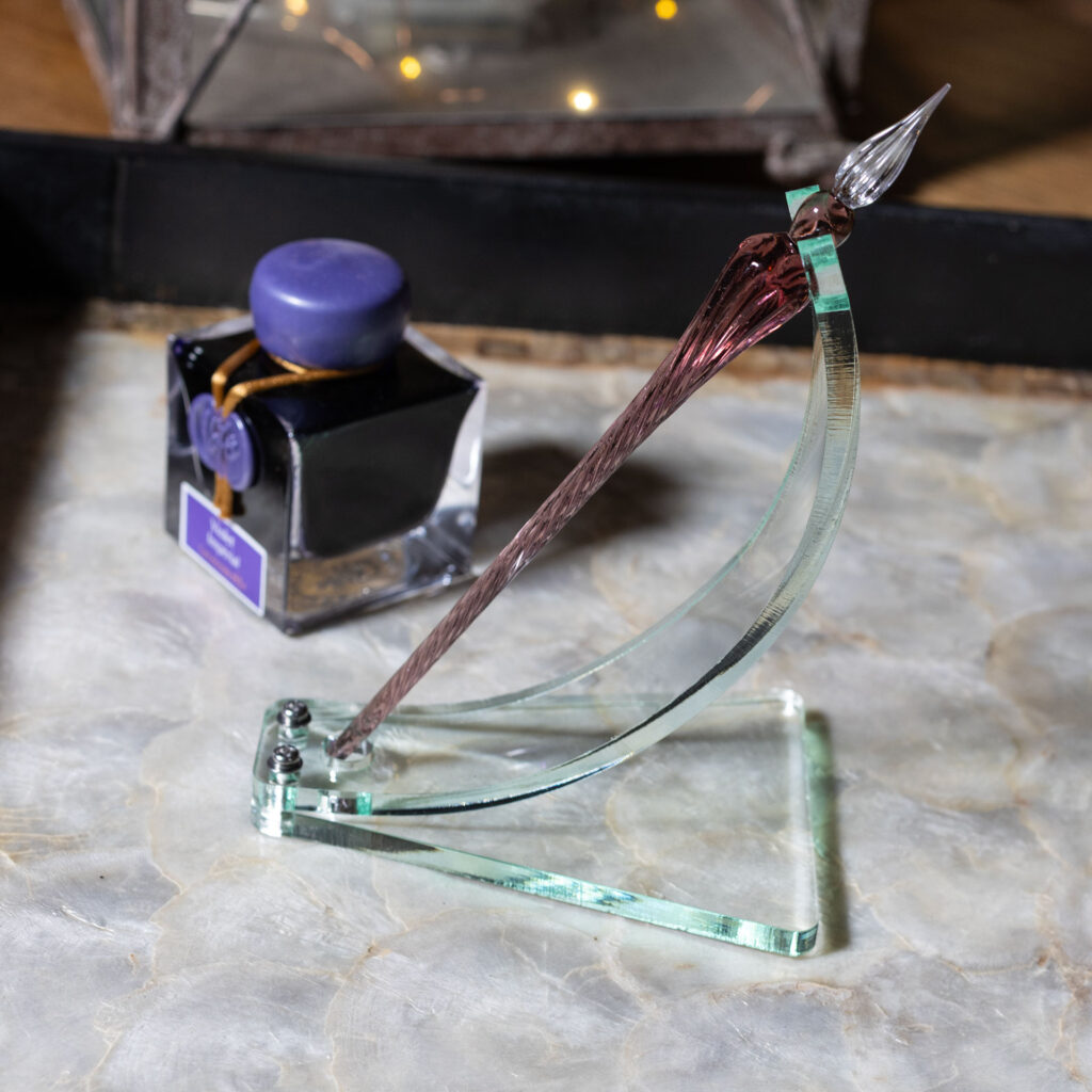 A green glass acrylic dip pen stand made with a curved strip bolted to a roughly trapezoidal base. A notch accommodates the shoulder of the pen at the top while a circular cut-out at the bottom keeps it from sliding. The elegran dip pen in the stand is violet with a clear tip. It sits next to a bottle of violet ink on a tray with a mother-of-pearl bottom and black sides.